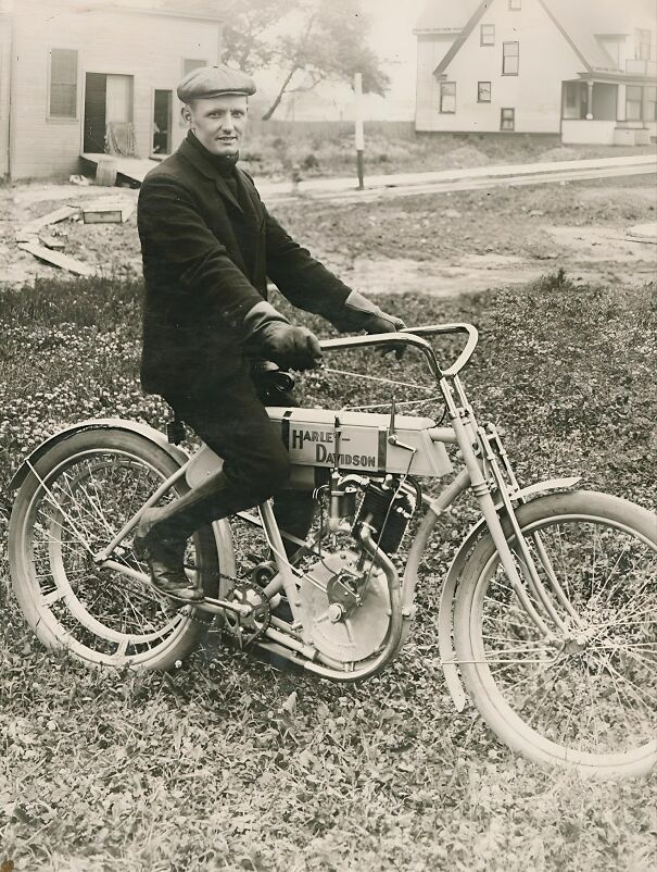 Man on vintage Harley-Davidson motorcycle, wearing a cap and gloves, in a historical rural setting.