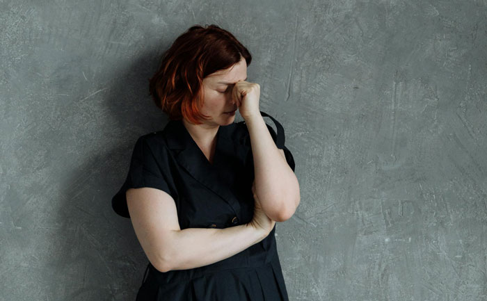 A woman in a black dress stands upset, covering her face against a gray wall. A woman in a black dress stands upset, covering her face against a gray wall.