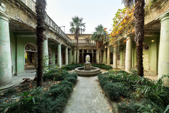 Abandoned place with decaying columns, a central fountain, overgrown plants, and an eerie, open courtyard.
