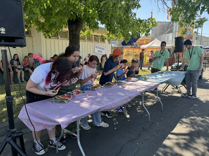 People participating in an outdoor eating competition, enjoying watermelon on a sunny day.