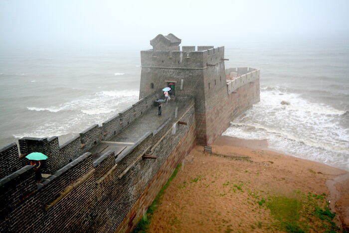 Great Wall of China at the sea's edge on a misty day, with people holding umbrellas.