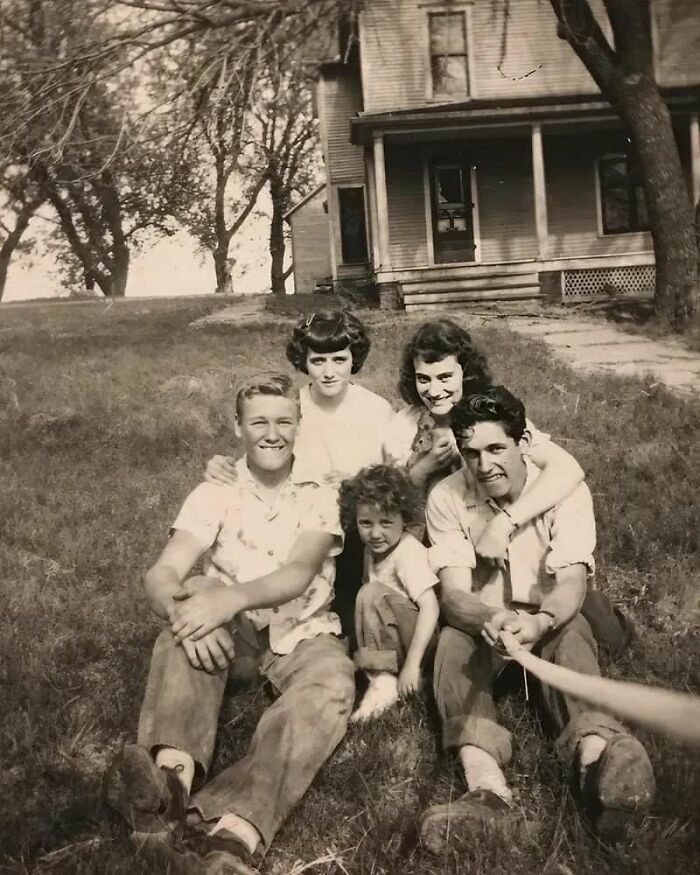 Vintage family snapshot sitting on grass, old house in background, capturing historical moments.