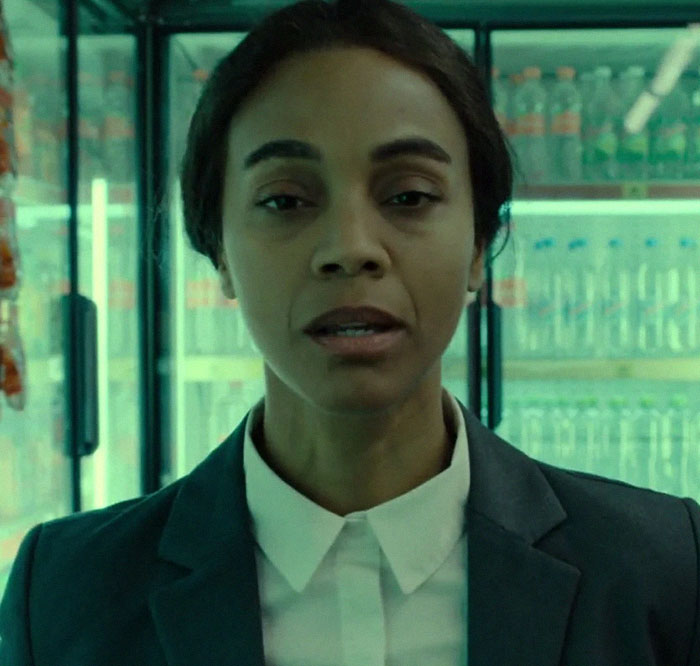A woman in a suit stands in front of a fridge with bottled drinks, looking serious.