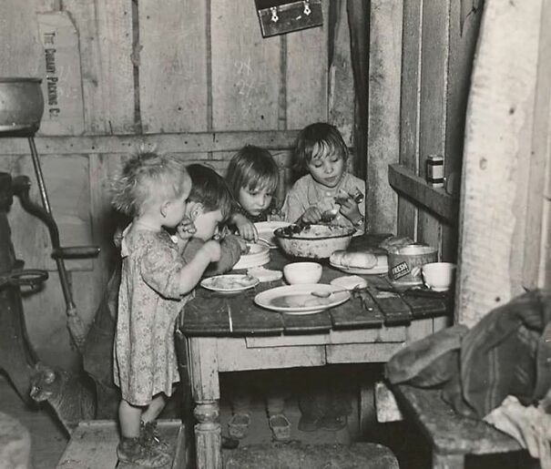 Four children in a rustic room, sharing a meal at a small table. Historical photo.