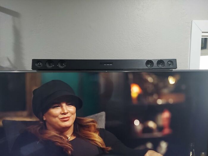 A black soundbar placed above a TV displaying a woman in a hat; ordinary setup with an extraordinary twist.