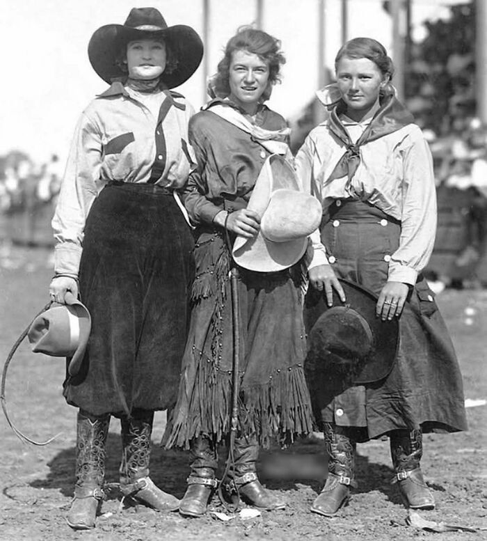 Historical photo of three women in cowboy attire, posing with hats and boots at a rodeo event.
