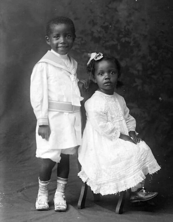 Two children in early 1900s attire, a boy in sailor suit and a girl in a lace dress, offer a glimpse into historical fashion.