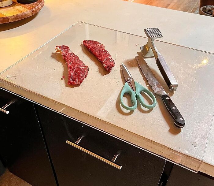 Kitchen tools, including scissors, a knife, and a meat tenderizer, alongside raw meat on a glass cutting board.