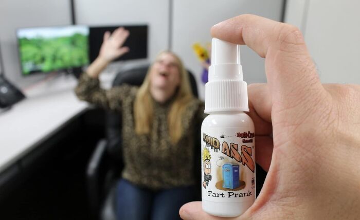 A prank product held in hand, with a blurred laughing woman in the office background.