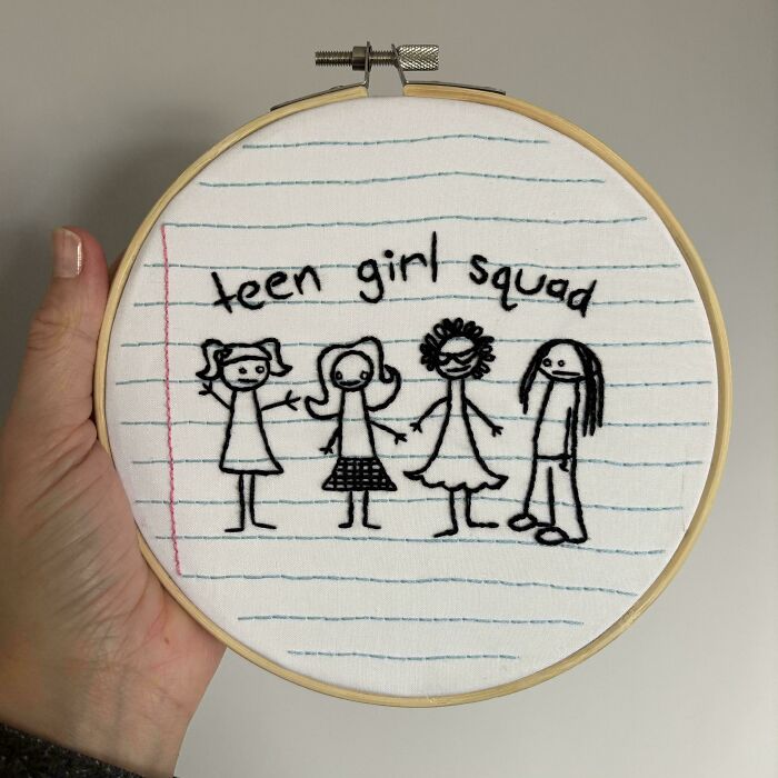 Hand holding embroidery hoop with stitched "teen girl squad" and stick figure girls on notebook paper fabric showcasing embroidery skills.