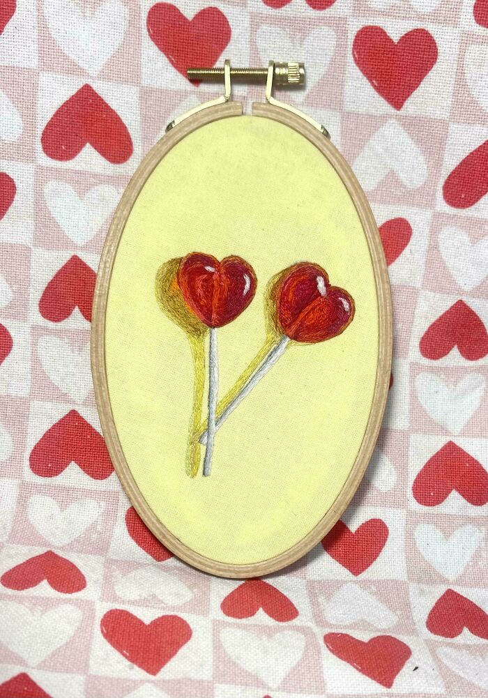 Embroidery hoop showing two realistic red heart-shaped lollipops stitched on yellow fabric with heart-patterned background.
