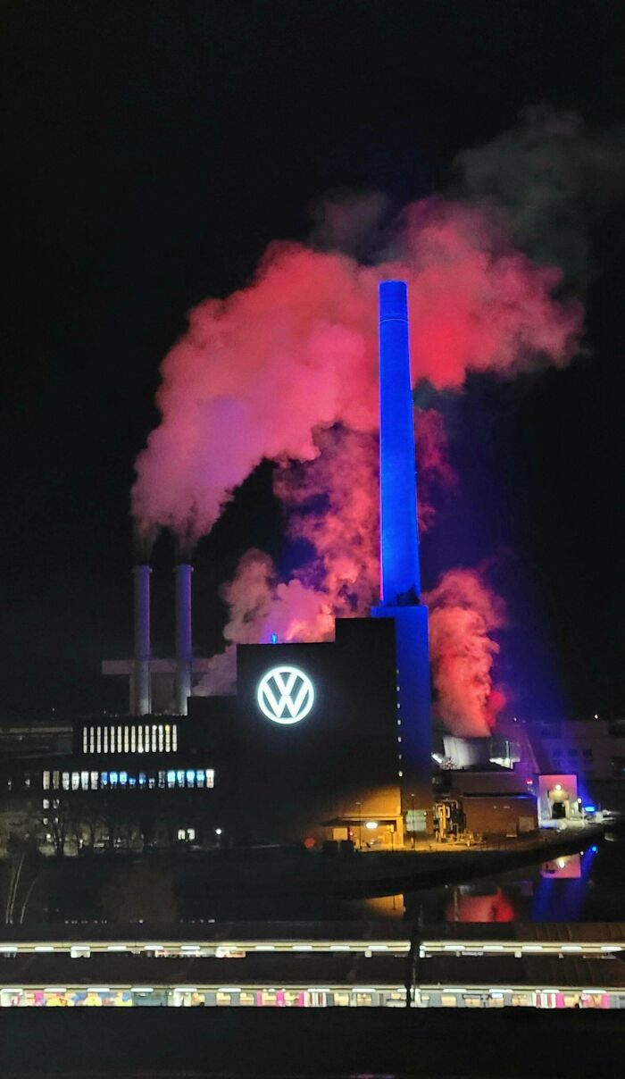 Industrial building glowing with red and blue lights at night, emitting smoke with an ominous supervillain headquarters vibe.