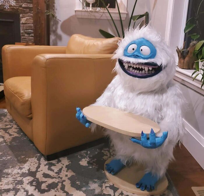 Creepy handmade craft of a white furry creature with blue face and hands, holding a round tray, next to a tan couch.
