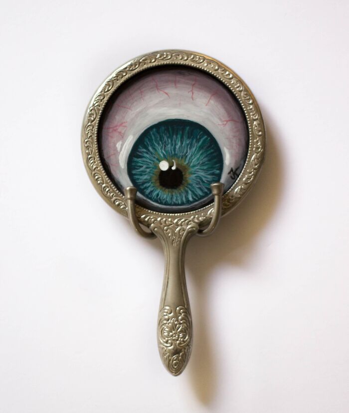 A silver vintage hand mirror with a painted eye in the center, representing creepy handmade crafts.