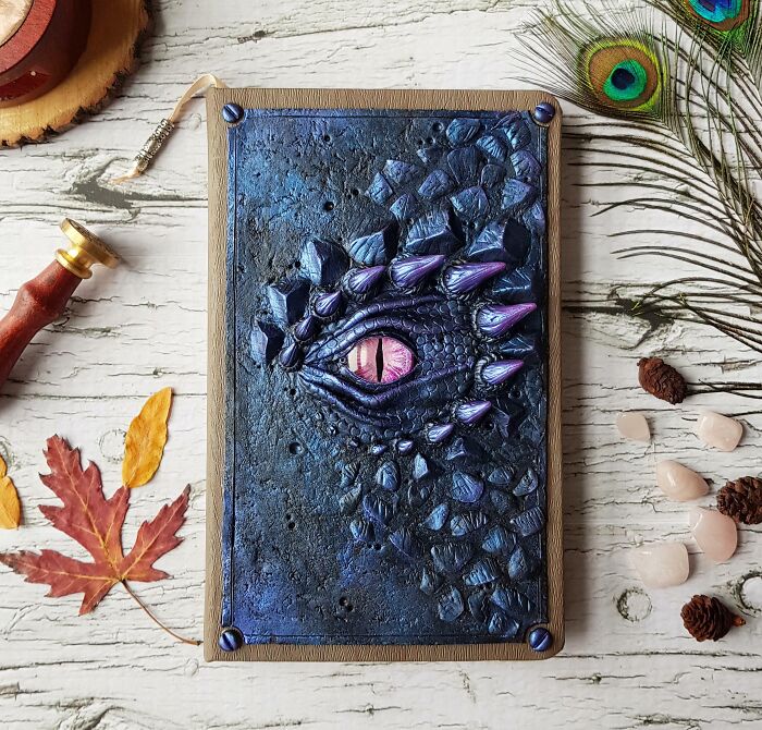 Creepy handmade craft featuring a dragon eye on a textured notebook cover, surrounded by leaves and stones.