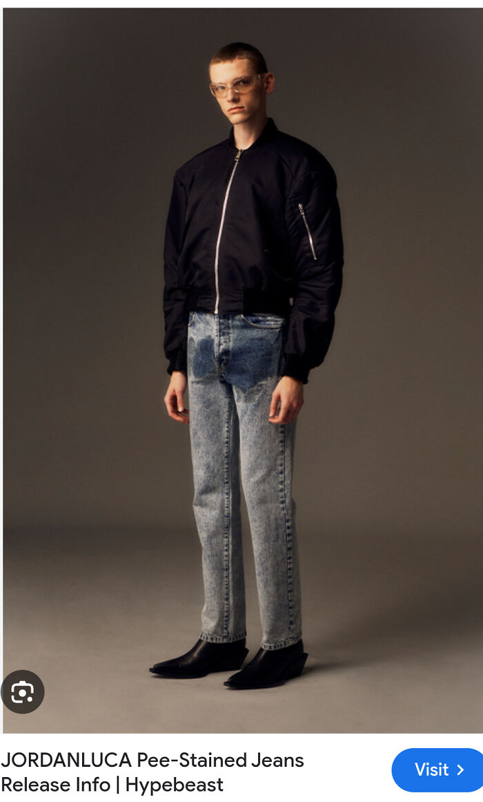 Hombre con chaqueta negra y jeans manchados, posando de pie.