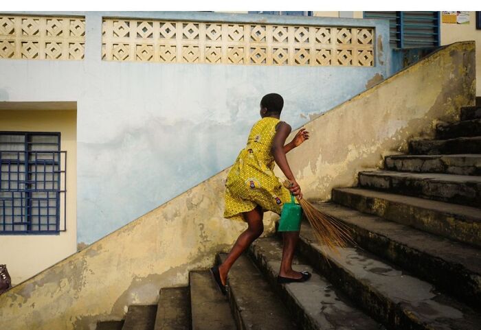 Andrea Torrei street photo: a person in a yellow dress climbing stairs, holding a broom and a green container, in an urban setting.
