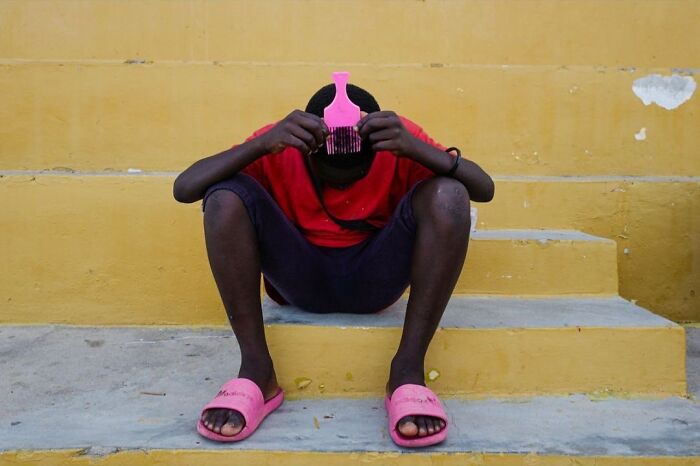 Person sitting on yellow steps, wearing a red shirt and pink slippers, using a comb. Street photo by Andrea Torrei.