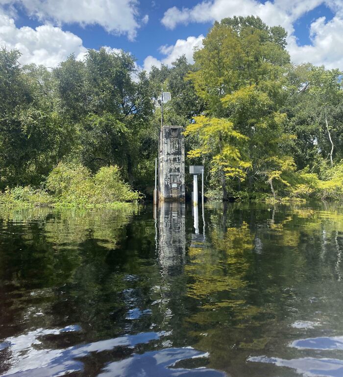 Weird object beside a lake surrounded by trees reflecting in the water under a partly cloudy sky.