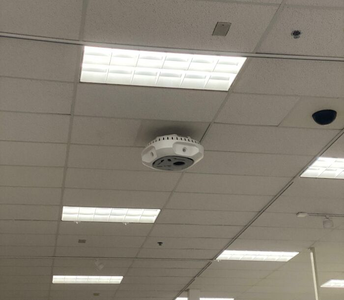Weird object on ceiling panel, possibly a sensor or camera, in a room with fluorescent lights.