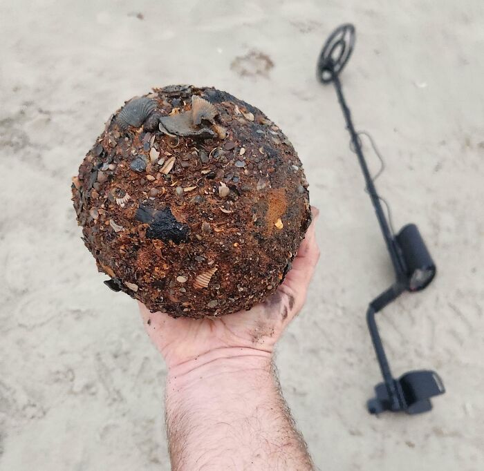 Hand holding a sand-covered, spherical object with a metal detector nearby on the beach.