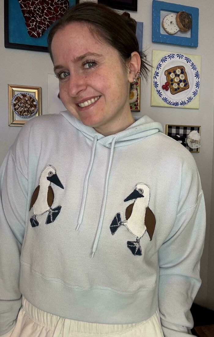 Person wearing a DIY sweatshirt with bird embroidery, smiling in front of decorative art pieces.