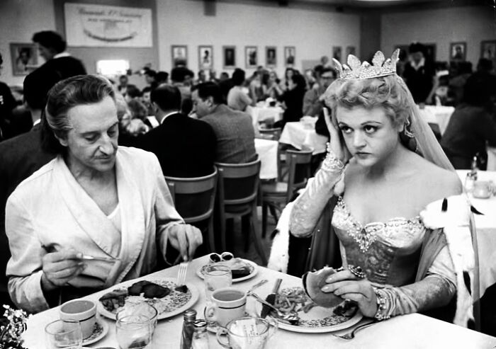 A vintage photo of two people dining in elegant attire, one wearing a crown, in a mid-20th century setting.