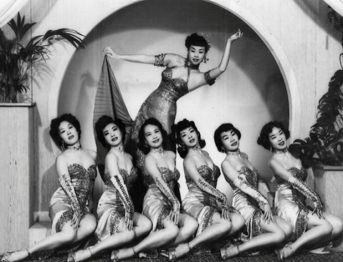 Vintage photo of seven women in ornate dresses striking a pose, showcasing timeless elegance from more than a hundred years ago.
