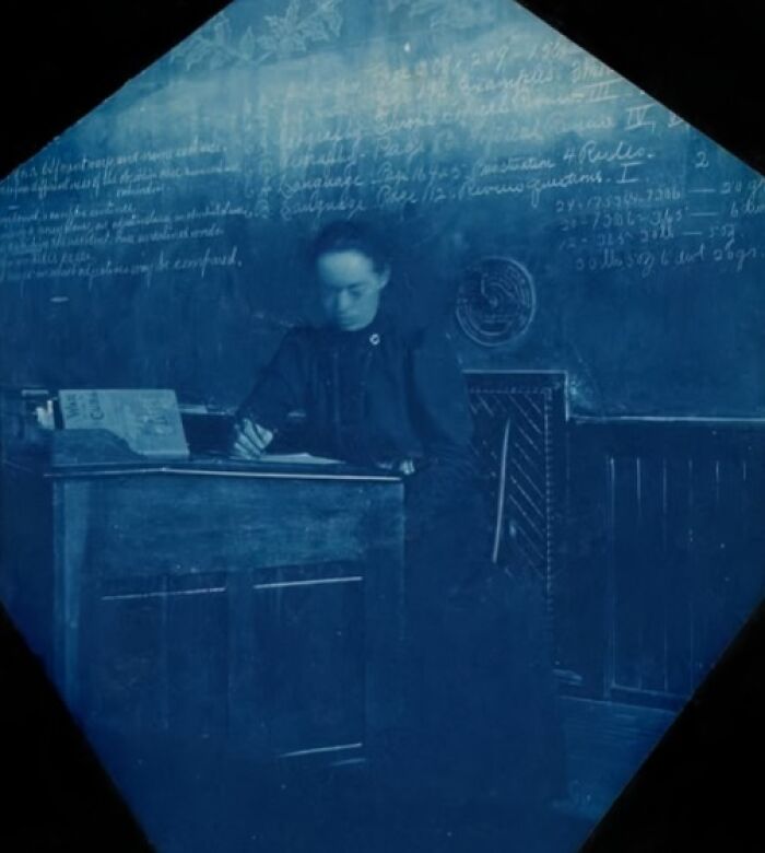 Woman in a classroom from a hundred years ago, writing at a desk, with a chalkboard covered in notes behind her.