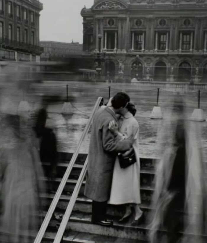 Couple kissing on stairs in historic black-and-white photo, with blurred pedestrians passing by.
