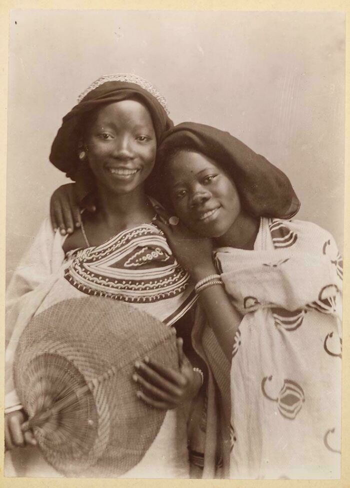 Two smiling women in traditional African attire and headwraps share a candid moment in a vintage historical photograph.