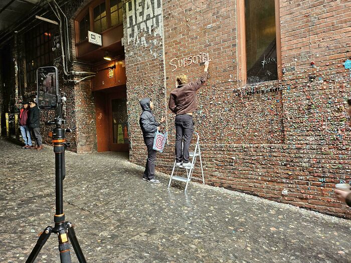 Person on ladder writing "Subscribe" on a gum-covered wall, capturing entitled behavior in urban setting.