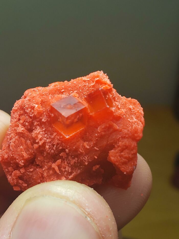 Red cube-shaped crystal formation held between fingers, showcasing natural things versus synthetic time effects.