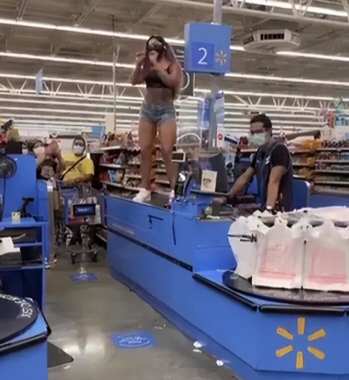 Woman standing on checkout counter, causing a scene in a grocery store, with masked employees and shoppers around.