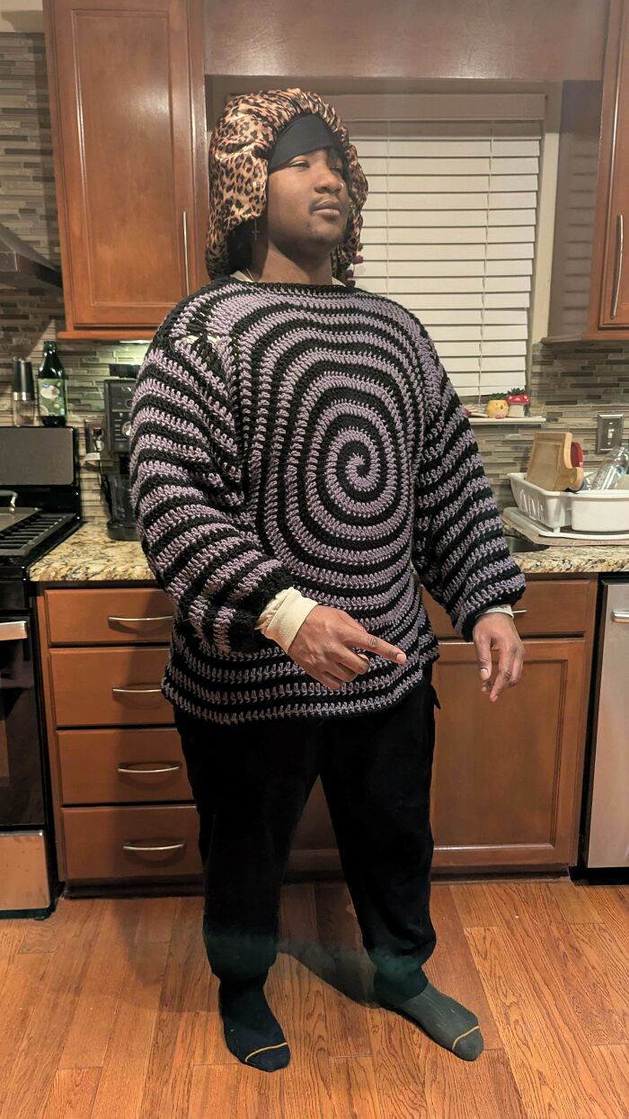 Man wearing a handmade crochet sweater with spiral pattern standing in a kitchen showcasing crochet creativity.
