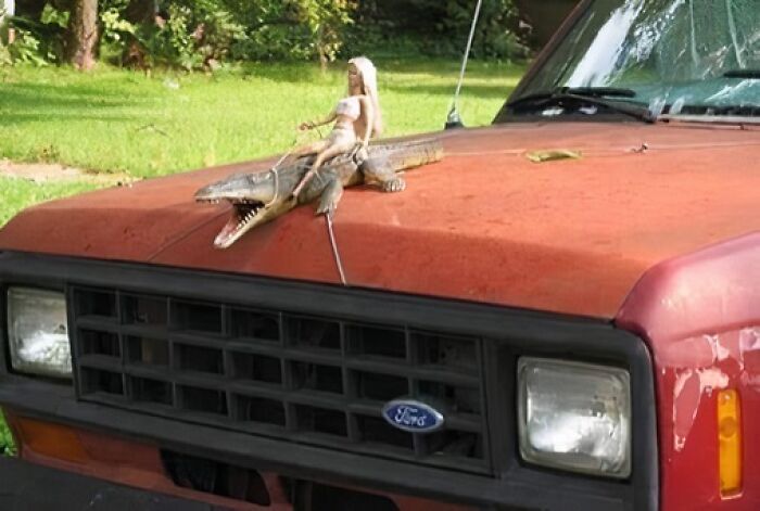 A Barbie doll riding a toy alligator on the hood of an old red Ford truck, showcasing random humor.