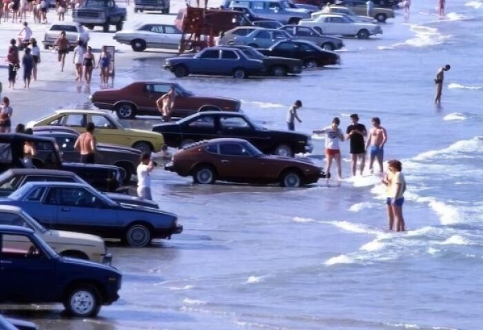 Cars parked on a crowded beach with waves reaching them, creating a funny random scene.