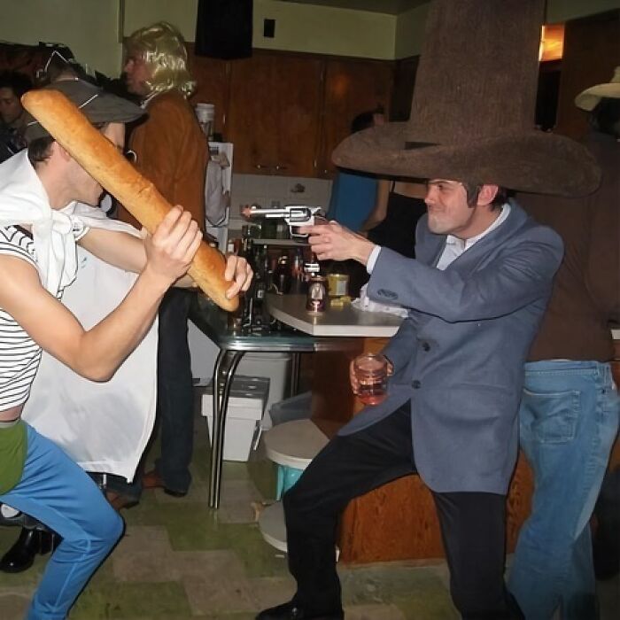 Two men in costumes having a funny standoff; one wields a baguette, the other a prop gun, at a party.