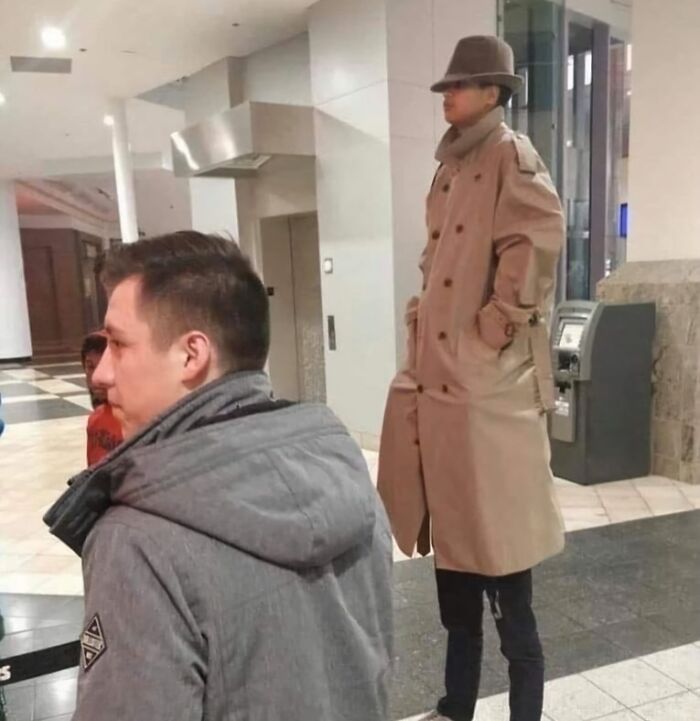 Person in a trench coat and hat standing on someone’s shoulders, looking humorous in a public space.