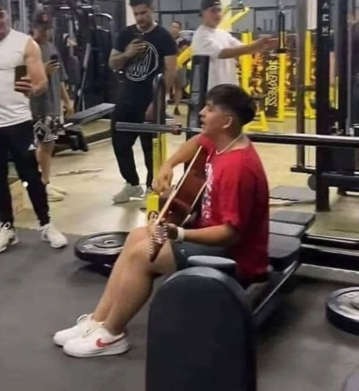 Man playing guitar in a gym while others watch, creating a funny random moment.