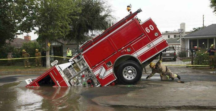 A fire truck is tilted into a flooded street, an accident or fail. Firefighters are present, surveying the expensive damage.