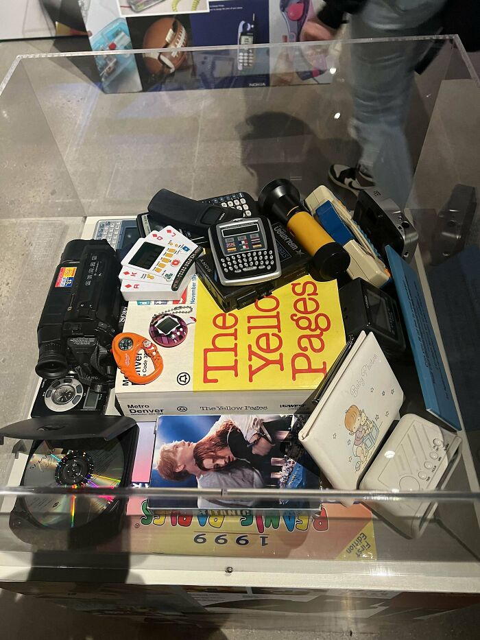 Collection of nostalgic 90s gadgets and items including a Yellow Pages book, video camera, and retro electronics in a display case.