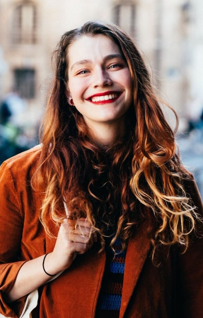 A woman with red lipstick and long hair smiles in a street portrait, wearing a brown jacket and holding a bag strap.