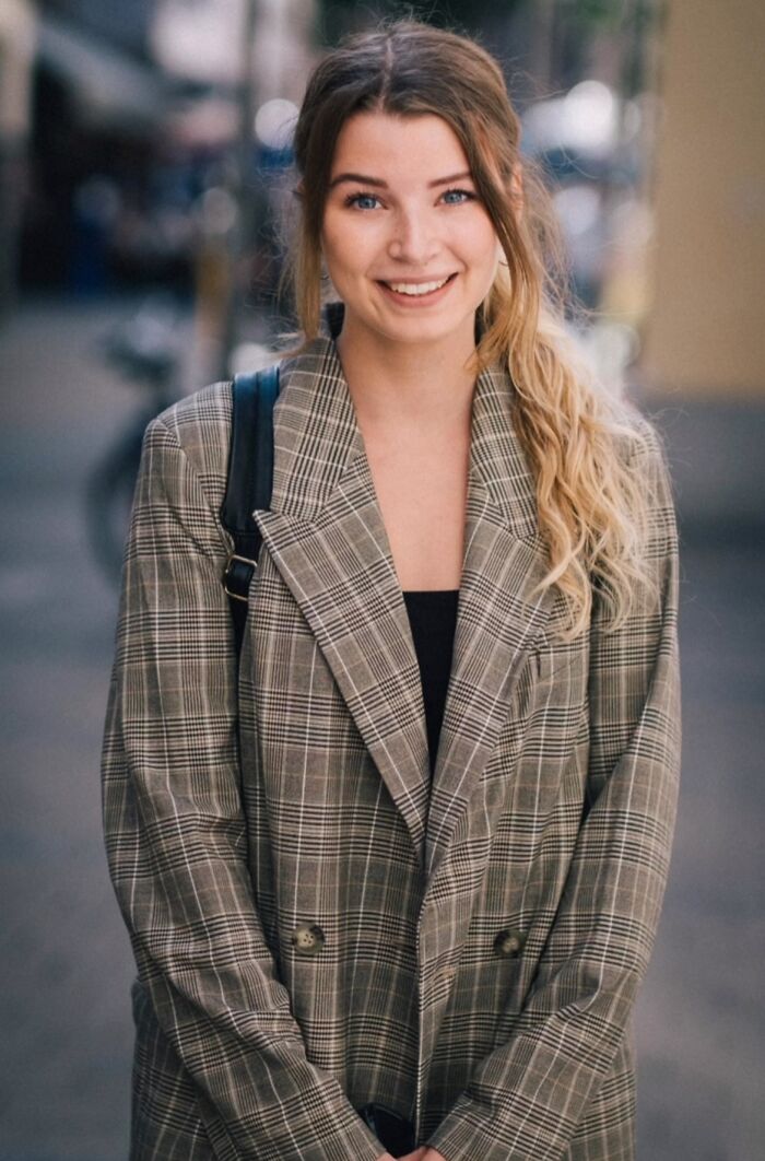 Street portrait of a smiling woman in a plaid coat, standing on an urban sidewalk.