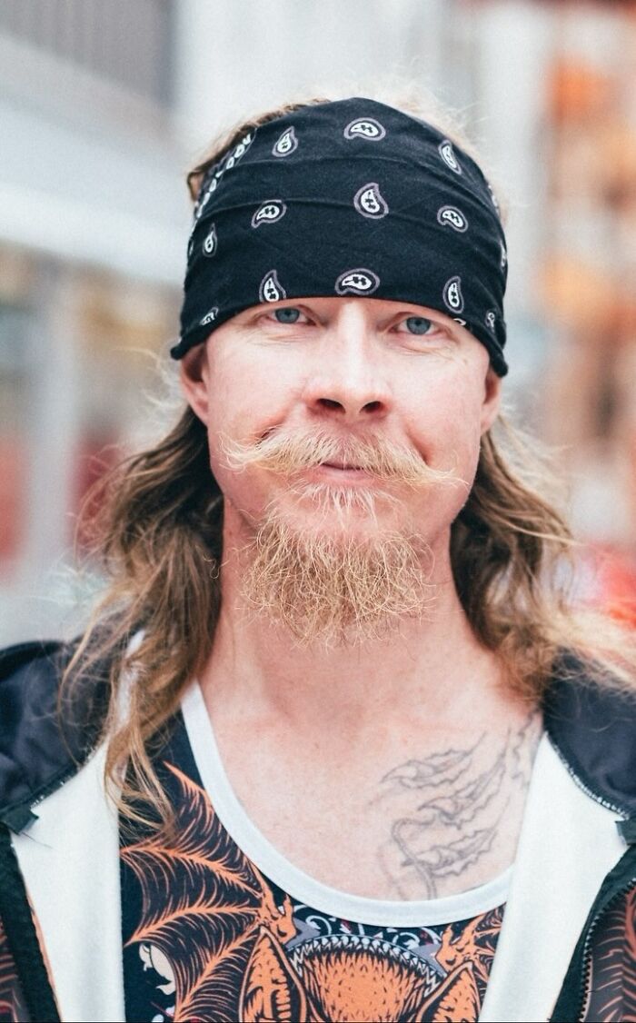 Long-haired man with a bandana, featuring a unique mustache and tattoo, as part of street portraits by a photographer.