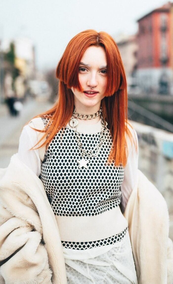 Red-haired woman in a fashionable street portrait wearing a patterned top, with blurred cityscape in the background.