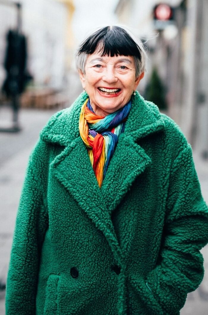 Elderly woman in a vibrant green coat and colorful scarf, smiling warmly in a street portrait.