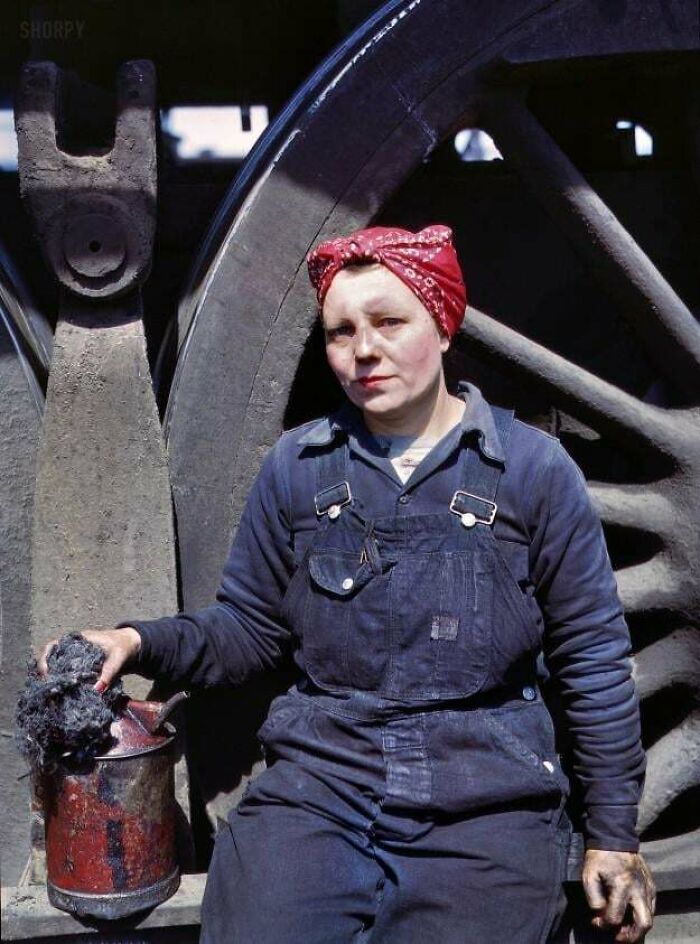 Woman worker in overalls with a red headscarf holding a coal bucket by large train wheels, candid glimpses into the past.