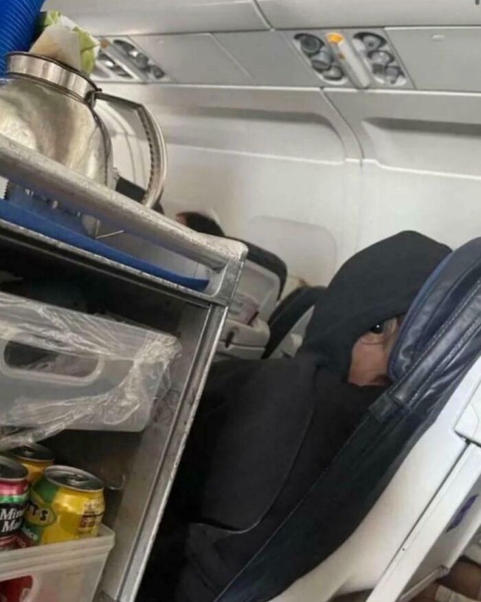 Confusing pic of a person in a hood peeking over an airplane seat near a beverage cart.