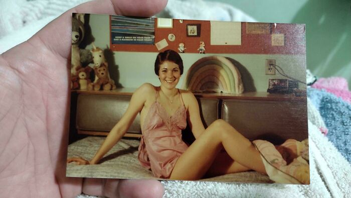 A hand holding a vintage photo of a woman sitting on a bed, surrounded by teddy bears and a rainbow decor.